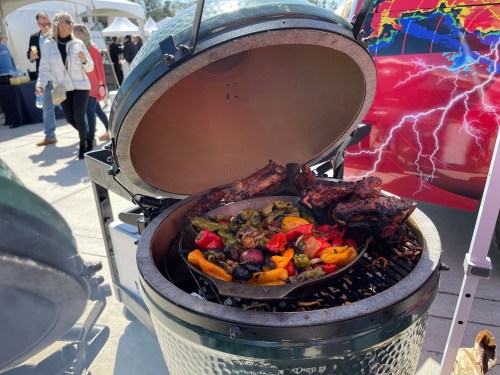 A grill with an open lid, featuring sizzling vegetables and meats, surrounded by a lively outdoor event.