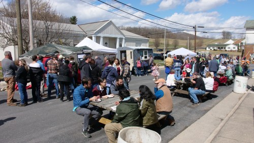 A bustling outdoor event with people eating at picnic tables and food vendors, set against a clear blue sky.