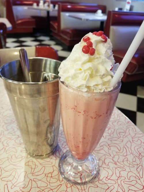 A tall glass of pink milkshake topped with whipped cream and red sprinkles, next to a metal cup on a retro diner table.