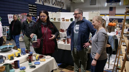 A diverse group of people examines handmade pottery at a craft fair in a gymnasium setting.