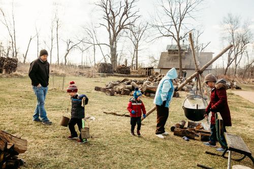 A group of children and an adult gather around a cooking pot outdoors, with firewood stacked in the background.