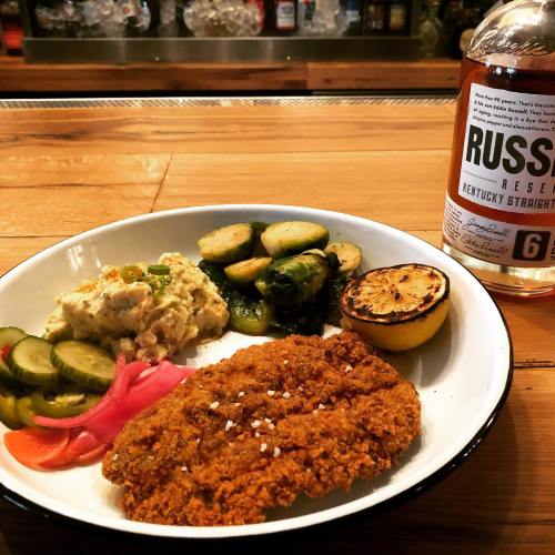 A plate with fried chicken, pickles, creamy salad, Brussels sprouts, grilled lemon, and a bottle of bourbon in the background.