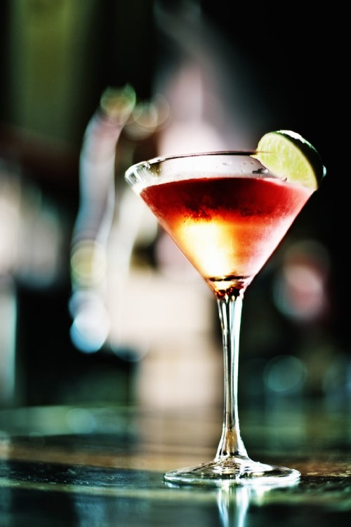 A vibrant cocktail in a martini glass, garnished with a lime slice, set against a blurred background.
