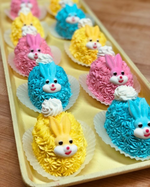 Colorful bunny-shaped cakes in pink, blue, and yellow, decorated with frosting and placed in a yellow tray.