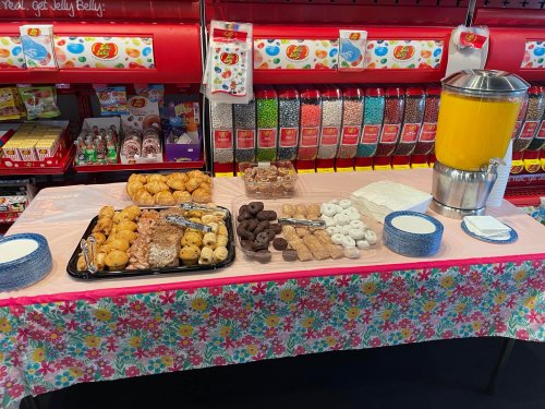 A table with various pastries, cookies, and a beverage dispenser filled with orange juice, set against a colorful backdrop.