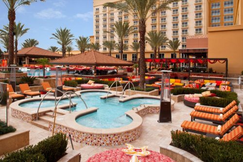 A vibrant pool area with lounge chairs, palm trees, and a hotel in the background, featuring hot tubs and colorful decor.
