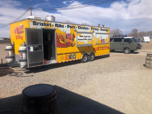 A bright yellow food truck named "Woody's Q Shack" parked in a gravel area, offering BBQ items like brisket and ribs.