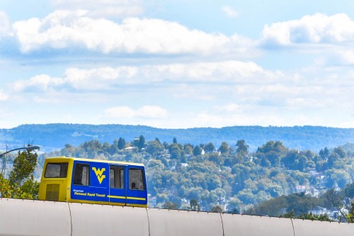 A blue and yellow monorail travels along an elevated track with a scenic view of hills and clouds in the background.