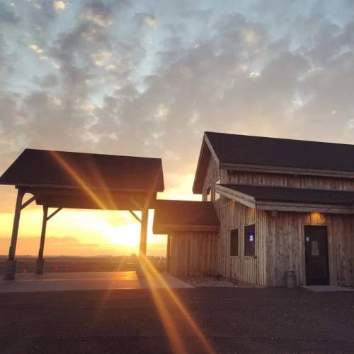 A wooden building with a covered area, illuminated by a sunset and surrounded by a cloudy sky.