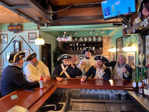 A group of six men in colonial attire sitting at a bar, enjoying drinks and chatting in a lively atmosphere.