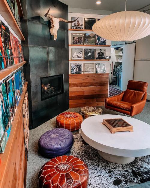 Cozy living room with a fireplace, leather chair, colorful poufs, and a round coffee table, featuring a unique wall decor.