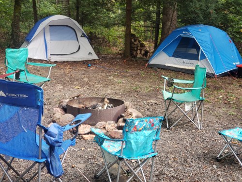 Two tents and several camping chairs surround a fire pit in a wooded area.