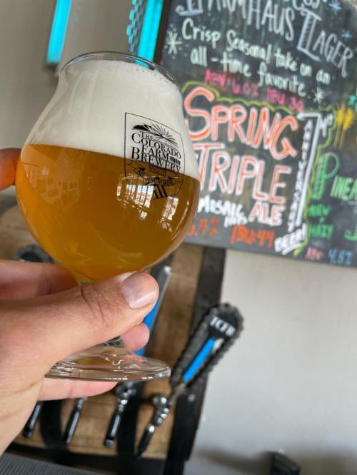 A hand holding a glass of light beer in front of a colorful chalkboard menu featuring various brews.