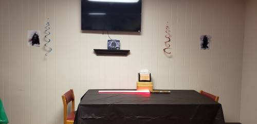 A table with a red lightsaber, a TV above, and decorations on the walls in a simple room setting.
