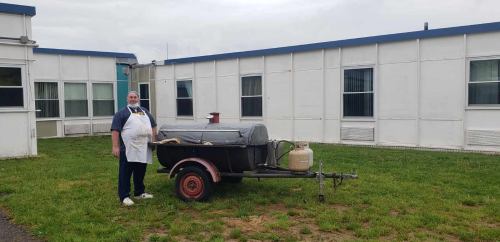 A person in an apron stands next to a trailer with a gas tank, set against a backdrop of modular buildings.