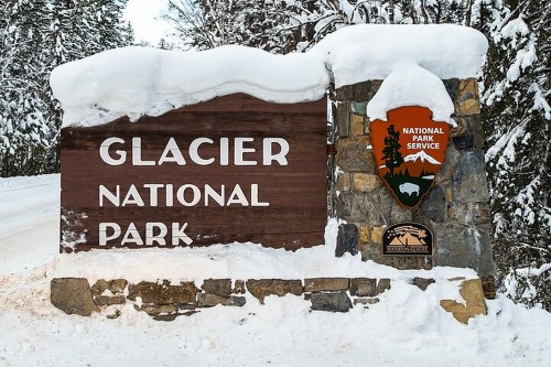 Sign for Glacier National Park covered in snow, with a National Park Service emblem beside it. Winter scenery.
