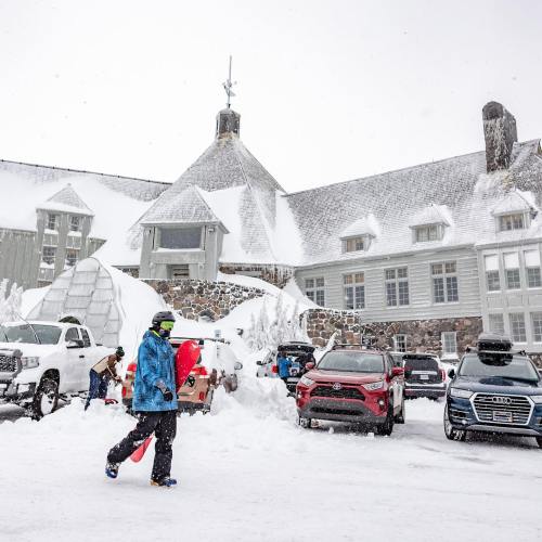 A snowy scene outside a lodge with people carrying snowboards and parked cars covered in snow.