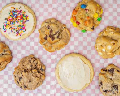 A variety of cookies on a pink checkered background, including frosted, chocolate chip, and colorful candy cookies.