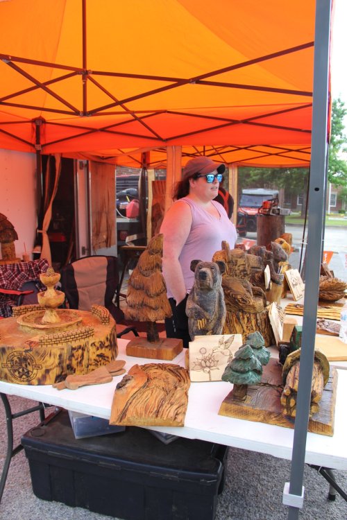 A woman stands behind a table displaying various wooden carvings under an orange tent at a market.