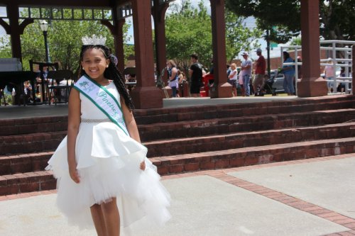 A young girl in a white dress and tiara smiles, standing in a park with people and a gazebo in the background.