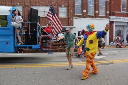 A colorful clown walks alongside a parade float with people, an American flag, and festive decorations in a lively street scene.