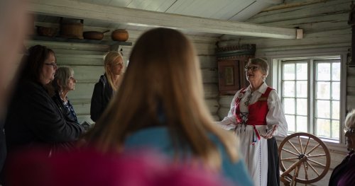 A group of people listens to a woman in traditional attire speaking inside a rustic cabin.