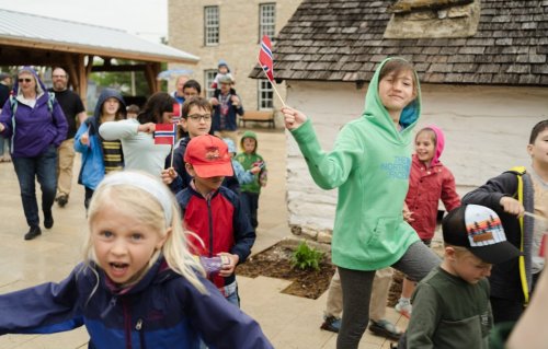 A group of children and adults joyfully parade outside, some waving flags, in a lively outdoor setting.