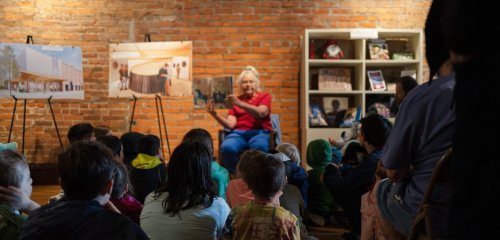 A woman sits in front of a group of children, sharing a story, with photos displayed on easels in a cozy setting.