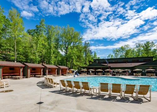 A sunny pool area surrounded by green trees, featuring lounge chairs and cabanas. Bright blue sky with clouds.