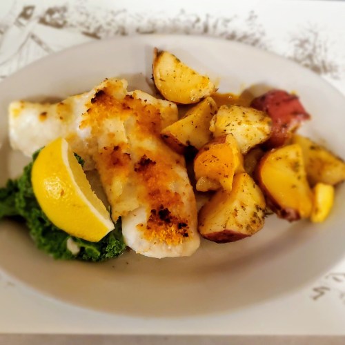 A plate of baked fish with golden crust, roasted potatoes, and a lemon wedge, garnished with greens.
