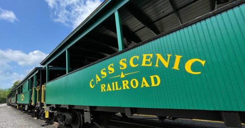A close-up of a green train car with "CASS SCENIC RAILROAD" painted in yellow on the side, under a blue sky.