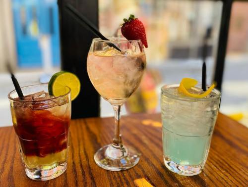 Three colorful cocktails on a wooden table: a dark drink with lime, a pink drink with a strawberry, and a light blue drink with lemon.