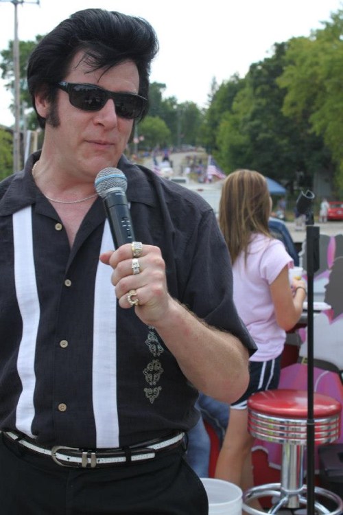 A man in sunglasses and a black shirt sings into a microphone at a parade, with a girl in the background.