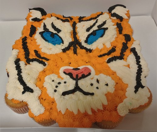 A cake decorated to resemble a tiger's face, featuring orange and white frosting with blue eyes and black details.