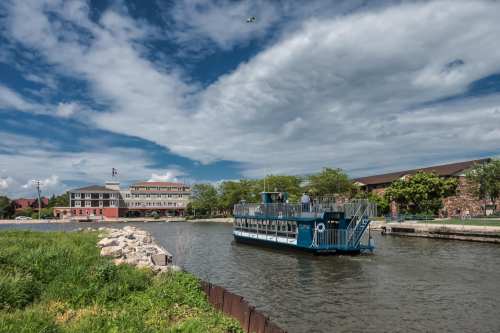 A blue boat navigates a calm waterway near a riverside building under a partly cloudy sky. Green grass lines the shore.