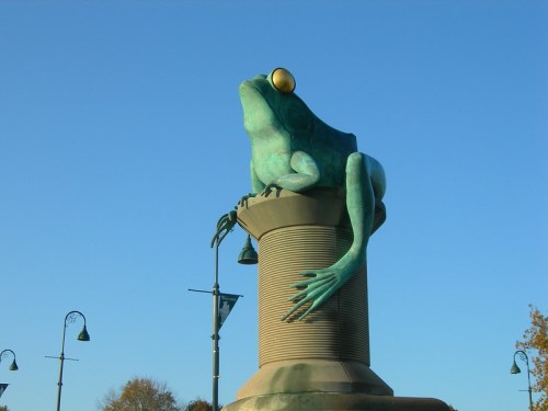 A large bronze frog sculpture perched on a cylindrical pedestal against a clear blue sky.