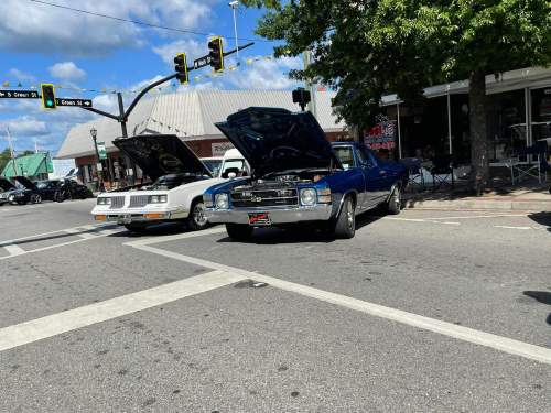 Two classic cars parked on a street with hoods open, under a blue sky with clouds and traffic lights in the background.