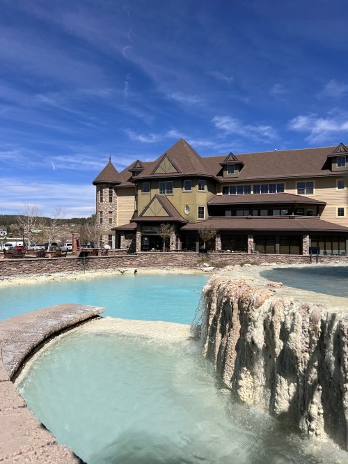 A large building with a stone facade beside a turquoise pool and waterfall under a blue sky with scattered clouds.