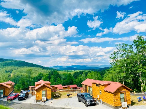 Scenic view of wooden cabins surrounded by lush greenery and mountains under a bright, cloudy sky.