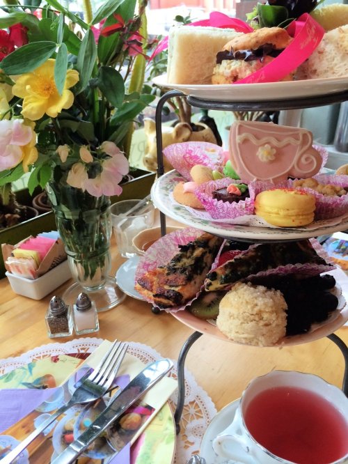A tiered stand of colorful pastries and treats beside a floral arrangement and a cup of tea on a wooden table.