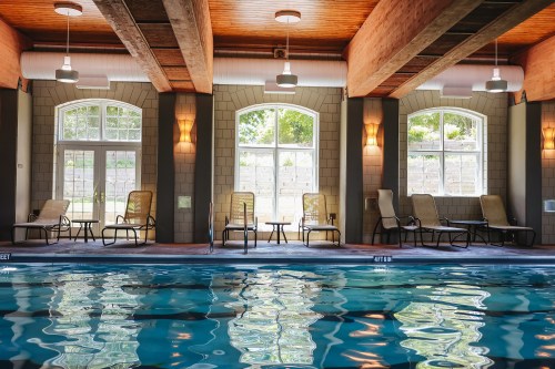 Indoor swimming pool with lounge chairs, large windows, and wooden beams overhead, reflecting light on the water.