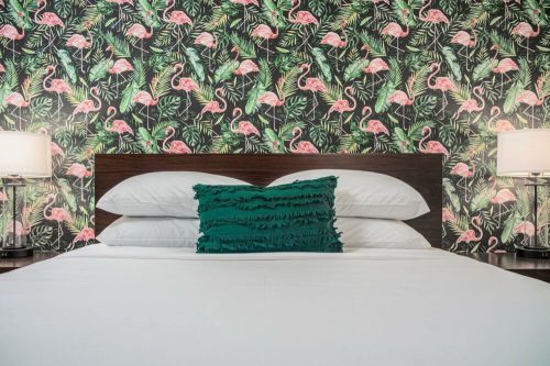 A bed with white linens and a teal decorative pillow, set against a vibrant flamingo-patterned wallpaper.