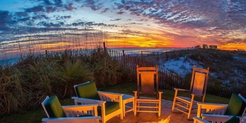 A serene beach scene at sunset, featuring a fire pit surrounded by chairs and a colorful sky.