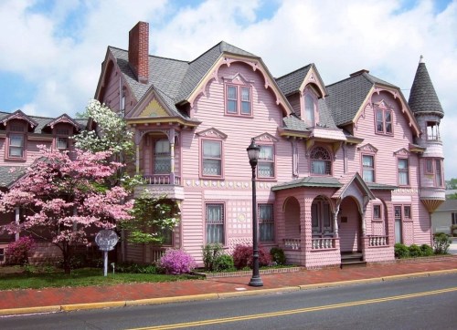 A large, ornate pink Victorian house with a turret, surrounded by blooming trees and flowers on a sunny day.