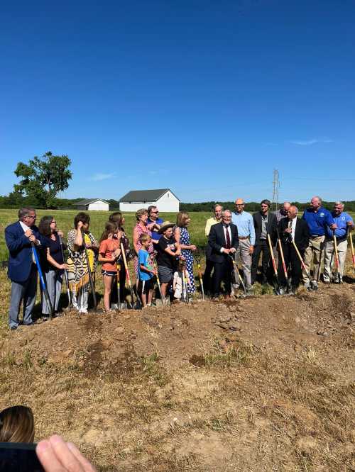 A group of people, including children, stand with shovels at a groundbreaking ceremony in a sunny outdoor setting.