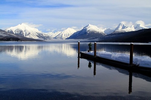 A serene lake surrounded by snow-capped mountains, with a person standing on a dock reflecting in the calm water.