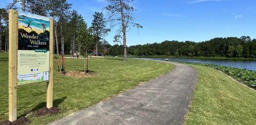 A scenic path by a lake, with a sign for "Wonder Walkers" and greenery surrounding the area.