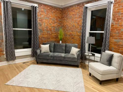 Cozy living room with exposed brick walls, a gray sofa, armchair, and a patterned rug. Natural light from large windows.
