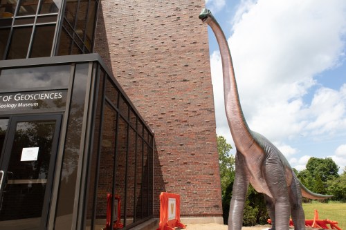 A tall dinosaur statue stands outside a brick building labeled "Department of Geosciences."