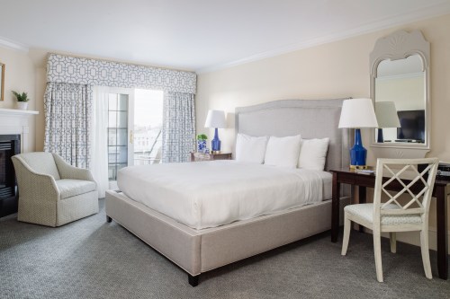 A cozy hotel room featuring a queen bed, desk, chair, and large windows with patterned curtains.